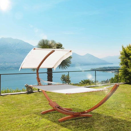 A stylish outdoor hammock with a wooden curved stand and a white canopy, set on a lush green lawn overlooking a serene lake and distant mountains under a clear blue sky. | PicSEO 1774622322378